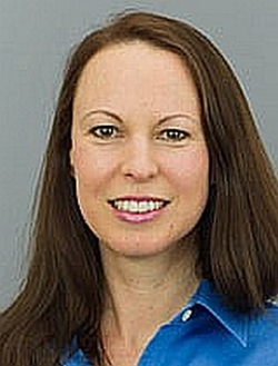 Kopfaufnahme einer lächelnden Frau mit langen braunen Haaren und blauem Hemd vor grauem Hintergrund.
