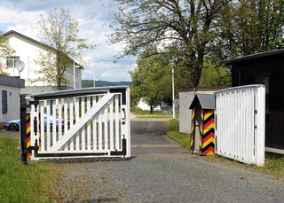 Offenes weißes Tor mit Deutschlandflaggenfarben an den Pfosten und einer gestreiften Wachkabine, führt zu einem gepflasterten Bereich.