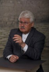 Ein älterer Mann mit grauem Haar, Brille und Schnurrbart in einem grauen Sakko, gestikulierend beim Sprechen.