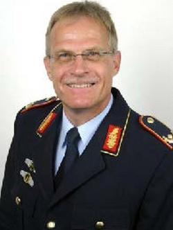 Lächelnder Mann mittleren Alters in dunkler Militäruniform mit Brille und Schnurrbart vor weißem Hintergrund.