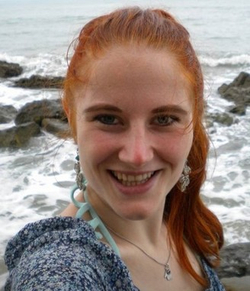 Lächelnde junge Frau mit roten Haaren und blauen Augen an einem felsigen Strand mit Wellen.