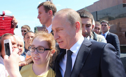 Wladimir Putin lächelt, während eine junge Frau ein Selfie mit ihm in einer Menschenmenge macht, mit weiteren Personen und Sicherheitspersonal.