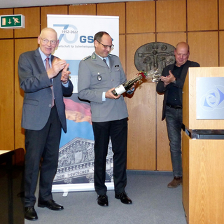 Drei Männer bei einer Veranstaltung. Ein Mann in Uniform hält eine Flasche und Rosen, während zwei weitere applaudieren.