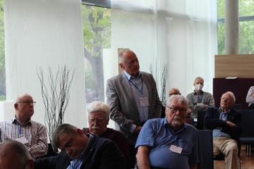 Ältere Männer bei einer Konferenz oder einem Treffen, ein Mann steht im grauen Anzug, andere sitzen, Fenster im Hintergrund.