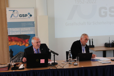 Zwei Männer an einem Konferenztisch mit Laptops und Mikrofonen, unter Bannern zum 70-jährigen Jubiläum der GSP.