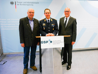 Drei lächelnde Männer, einer in Uniform, zwei in Zivil, vor einem Hintergrund des Presseamtes der Bundesregierung.