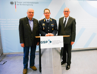 Drei lächelnde Männer, einer in Uniform, zwei in Zivil, vor einem Hintergrund des Presseamtes der Bundesregierung.