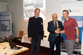 Drei Männer bei einer Veranstaltung. Zwei schütteln sich die Hände, einer hält eine kleine Flasche. Banner und eine Leinwand im Hintergrund.