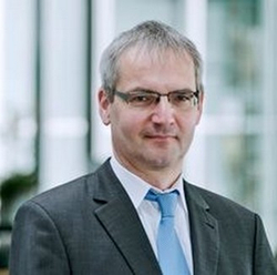 Porträt eines Mannes mit grauem Haar und Brille, Anzug und blauer Krawatte, blickt nach vorne.