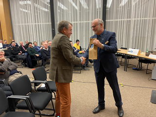 Zwei lächelnde Männer schütteln sich die Hände, einer hält eine GSP-Geschenktüte, bei einer Veranstaltung mit Publikum.