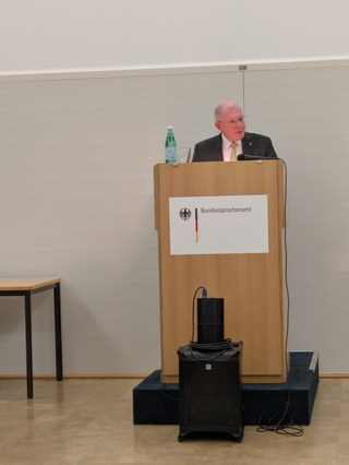 Ein älterer Mann in Anzug spricht an einem Rednerpult mit dem Logo des Bundessprachenamtes. Wasserflasche und Mikrofon sind zu sehen.