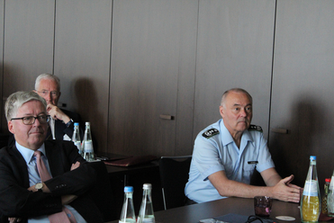 Drei Männer, zwei in Anzügen und einer in hellblauer Uniform mit Schulterklappen, sitzen am Konferenztisch.