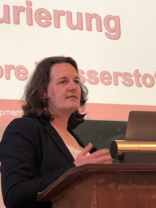 Eine Frau spricht an einem Rednerpult bei einer Präsentation, mit Teilen deutscher Wörter wie 'Wasserstoff' auf der Leinwand.