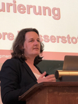 Eine Frau spricht an einem Rednerpult bei einer Präsentation, mit Teilen deutscher Wörter wie 'Wasserstoff' auf der Leinwand.