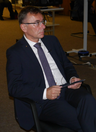 Ein Mann im dunklen Anzug mit Brille sitzt und hält ein Tablet. Er trägt ein Ansteckmikrofon und einen goldenen Pin.
