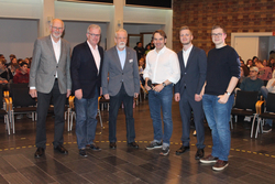 Sechs Männer in Business-Casual-Kleidung lächeln bei einem Treffen, einer hält ein Mikrofon, mit Publikum im Hintergrund.