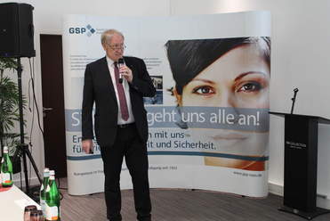 Mann im dunklen Anzug spricht in ein Mikrofon bei einer Veranstaltung, mit großem GSP-Banner und Frauengesicht dahinter.