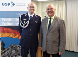Porträt von zwei lächelnden Männern, einem Uniformierten und einem im Sakko, vor einem Hintergrund mit GSP-Logo und Deutschlandflagge.