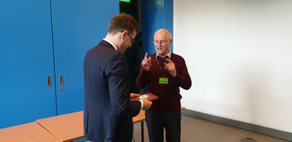 Ein Mann im Anzug erhält eine Plakette von einem älteren Mann mit „Tribüne“-Namensschild, der spricht und gestikuliert.