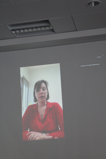 Projektion von Eva-Maria Steinbrecher, einer Frau in rotem Hemd und Brille, in einem Videoanruf, sprechend oder präsentierend.
