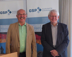 Zwei lächelnde Männer, möglicherweise von der Gesellschaft für Sicherheitspolitik (GSP), vor Veranstaltungsbannern.