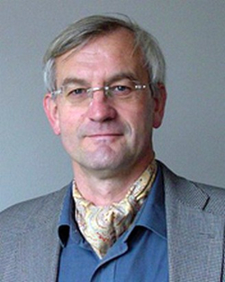 Kopfaufnahme eines Mannes mit grauem Haar, Brille, blauem Hemd, grauer Jacke und gemustertem Halstuch.