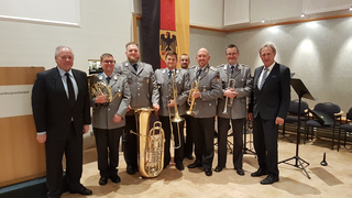 Mitglieder einer Bundeswehr-Militärkapelle mit Blechblasinstrumenten und zwei Zivilisten vor einer Deutschlandflagge.