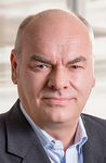 Ein glatzköpfiger Mann mit blauen Augen lächelt leicht, trägt eine dunkle Anzugjacke und ein hellblaues gestreiftes Hemd.