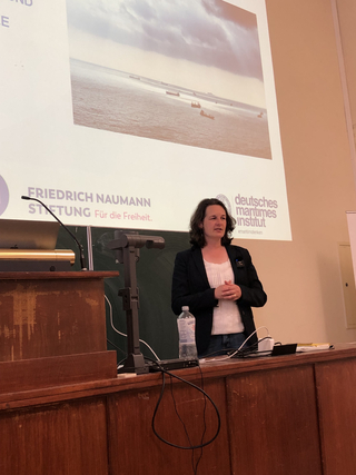 Frau präsentiert an einem Rednerpult; Leinwand zeigt Schiffe und Logos der Friedrich Naumann Stiftung & Deutsches Maritimes Institut.