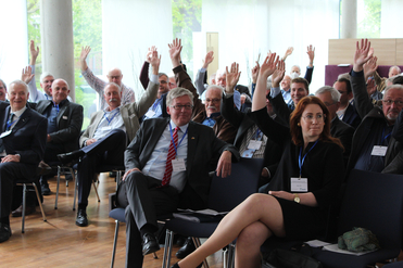 Eine Gruppe von Konferenzteilnehmern, darunter Männer und eine Frau, sitzt und hebt die Hände zum Abstimmen oder Fragenstellen.