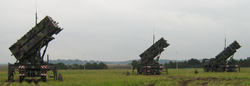 Drei Patriot-Raketenwerfer, schräg nach oben gerichtet, sind auf einem Grasfeld mit Soldaten unter bewölktem Himmel stationiert.