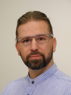 Porträt eines Mannes mit zurückgebundenem Haar, Brille, braunem Bart und blau-weiß gestreiftem Hemd.