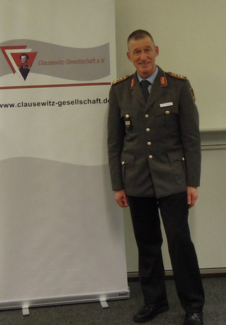 Ein lächelnder Mann in dunkler Militäruniform steht neben einem Banner der Clausewitz-Gesellschaft e.V.