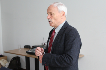 Älterer Mann mit weißem Haar und Schnurrbart, in Sakko und roter Krawatte, sprechend und seine Manschette richtend.
