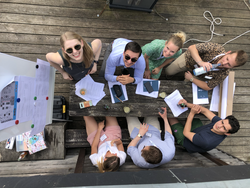 Gruppe junger Leute an einem Holztisch im Freien, lächelnd in die Kamera blickend. Papiere und Getränke liegen auf dem Tisch.