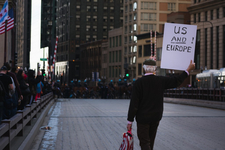 Ein Mann mit einem Schild 'US AND ! EUROPE' geht auf einer Stadtstraße mit Gebäuden und Polizei im Hintergrund.