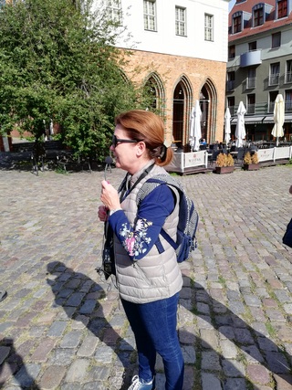 Eine Frau mit Mikrofon und Sonnenbrille auf einer Kopfsteinpflasterstraße vor historischen Gebäuden und einem Café.
