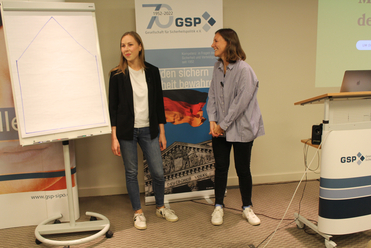 Zwei Frauen, eine lächelnd und eine lachend, stehen vor GSP-Bannern und einem Flipchart während einer Präsentation.
