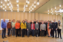 Eine große Gruppe meist älterer Erwachsener, darunter ein Mann in Uniform, posiert für ein Gruppenfoto in einem hellen Saal.