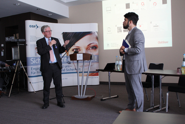 Ein Mann spricht ins Mikrofon während einer Konferenz; ein anderer Mann hört zu. Eine Projektion und ein Banner im Hintergrund.