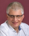 Lächelnder Mann mit kurzen grauen Haaren und Brille im weißen Hemd.