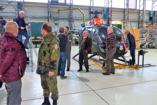 Mehrere Personen, darunter Militärangehörige, versammeln sich um einen dunklen Hubschrauber in einem Hangar.