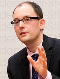 Ein Mann mit Brille, dunklem Anzug, rosa Hemd und gepunkteter Krawatte, gestikuliert mit erhobener Hand.