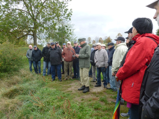Gruppe älterer Menschen, darunter ein sprechender Mann und eine Frau mit buntem Regenschirm, versammelt sich im Freien auf einer Wiese.