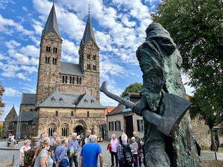 Bronzestatue einer bekleideten Figur mit Axt, eine Reisegruppe und eine große Steinkirche mit zwei Türmen vor bewölktem Himmel.