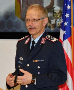 Ein deutscher Militäroffizier in Uniform, mit Brille und Schnurrbart, steht vor einer amerikanischen Flagge.