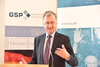 Ein Mann im Anzug spricht an einem Rednerpult und gestikuliert. GSP- und KAS-Banner im Hintergrund.