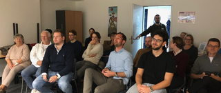 Eine vielfältige Gruppe von Menschen sitzt aufmerksam lauschend in einem hellen Raum, einige lächeln.