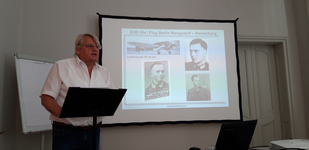 Ein Mann präsentiert eine historische Bilderschau mit Flugzeug, Soldatenporträts (inkl. Stauffenberg) und Flugdetails.