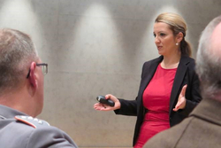 Eine Frau in rotem Kleid und schwarzem Blazer präsentiert mit einem Klicker vor einem Publikum, darunter ein Militärangehöriger.